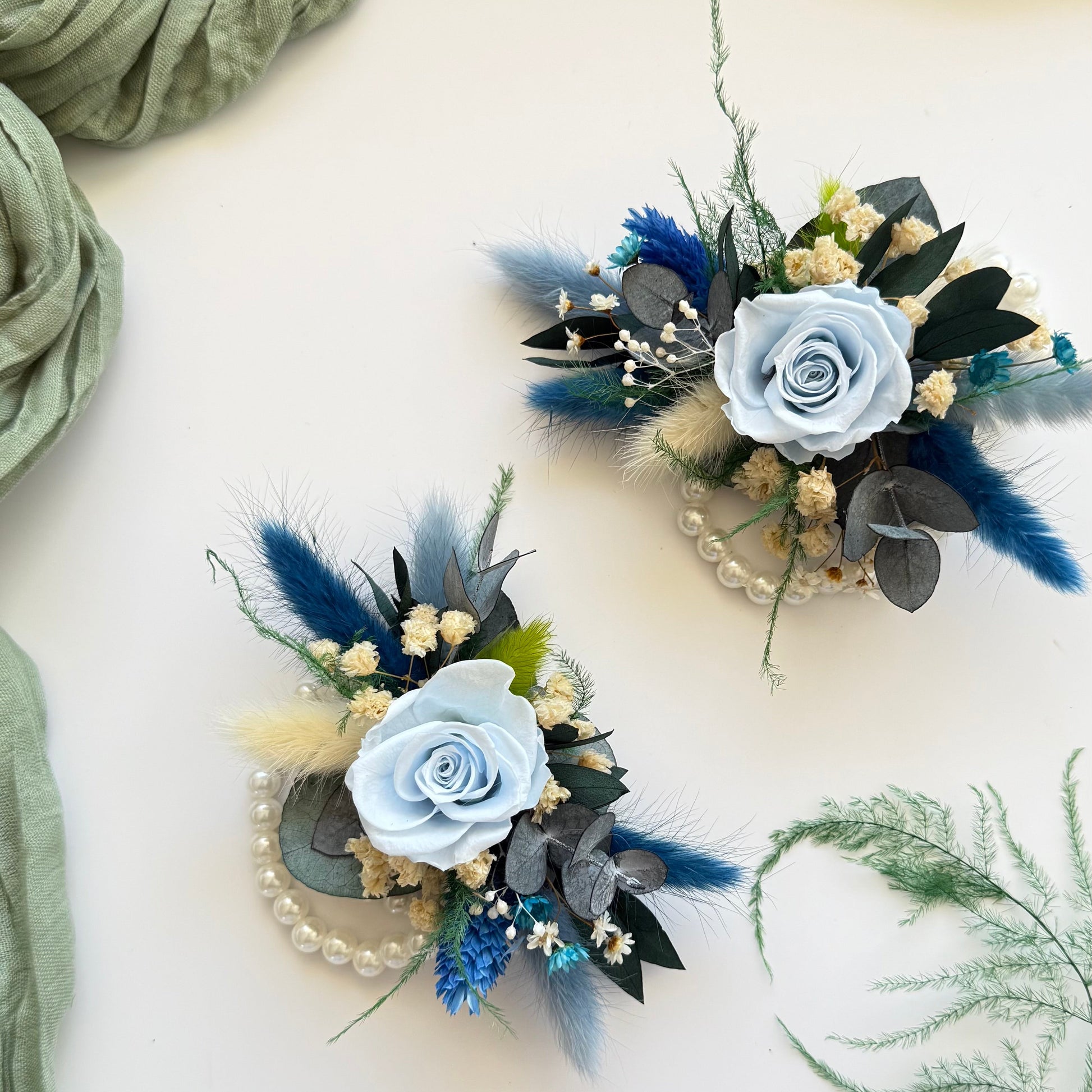 Two floral corsages with blue flowers and greenery on a light background