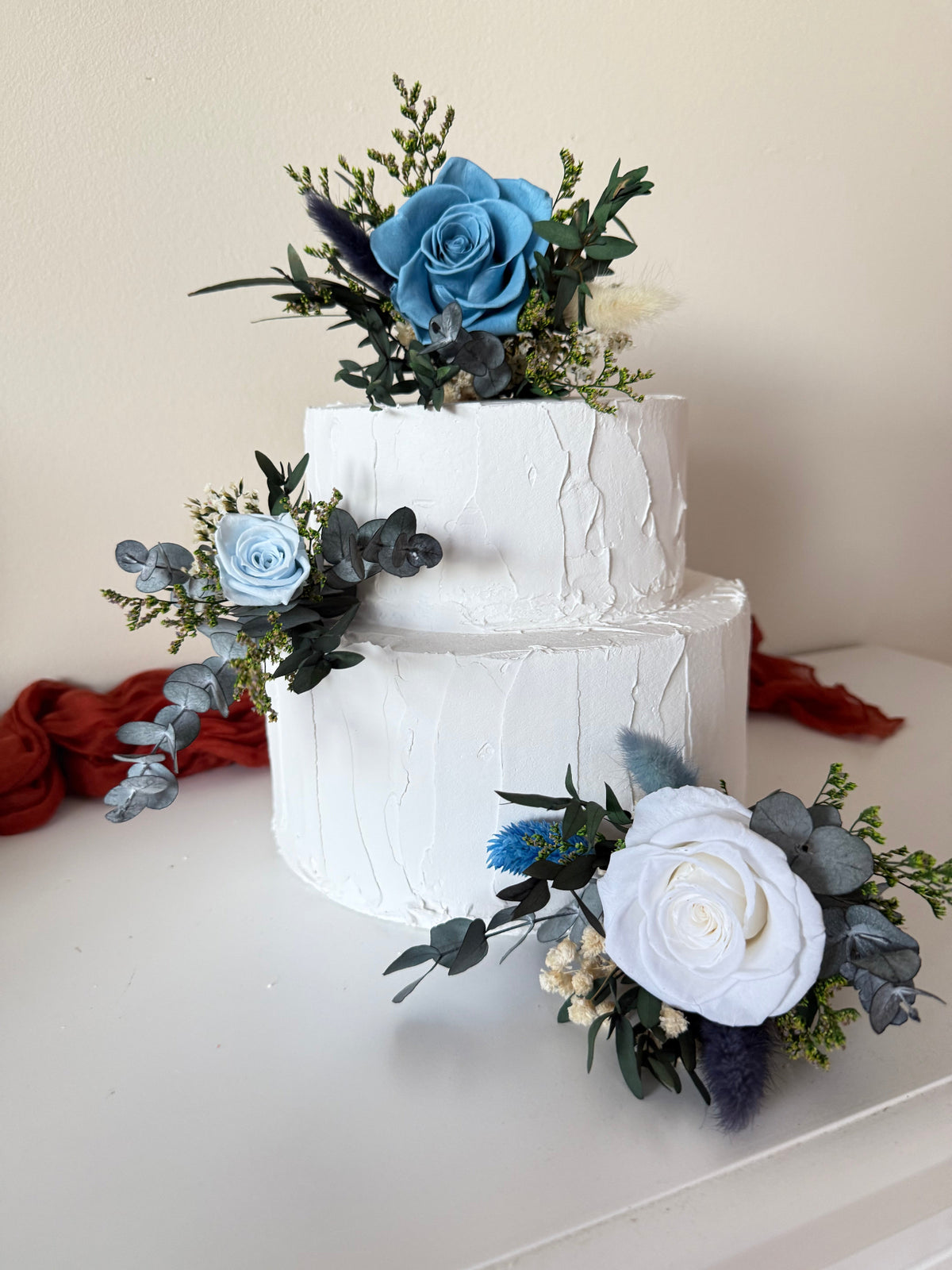 Elegant wedding cake styling with soft blue roses, ivory flowers and natural preserved greenery in a rustic, modern botanical palette.