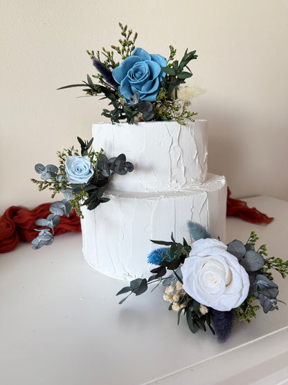 Elegant wedding cake styling with soft blue roses, ivory flowers and natural preserved greenery in a rustic, modern botanical palette.
