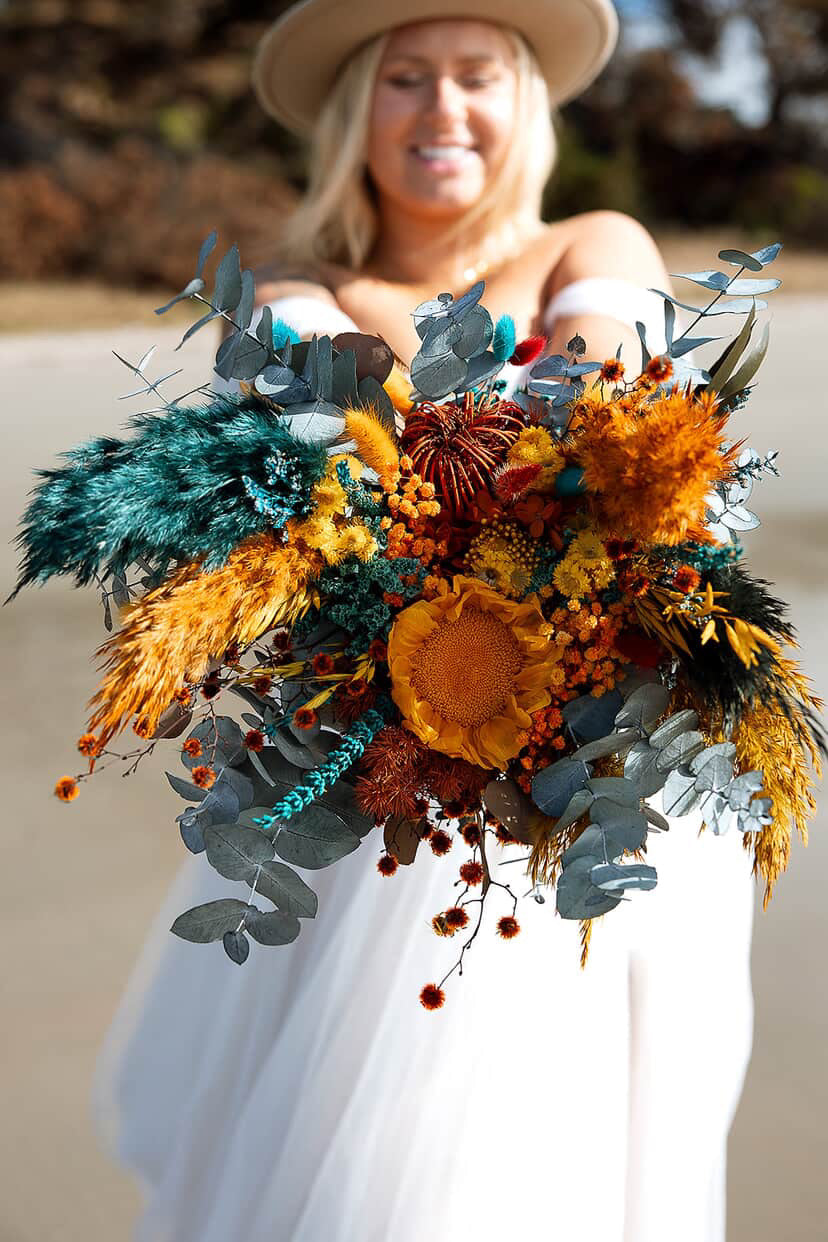 Bride holding sunflower and teal dried flower wedding bouquet with preserved eucalyptus and burnt orange accents