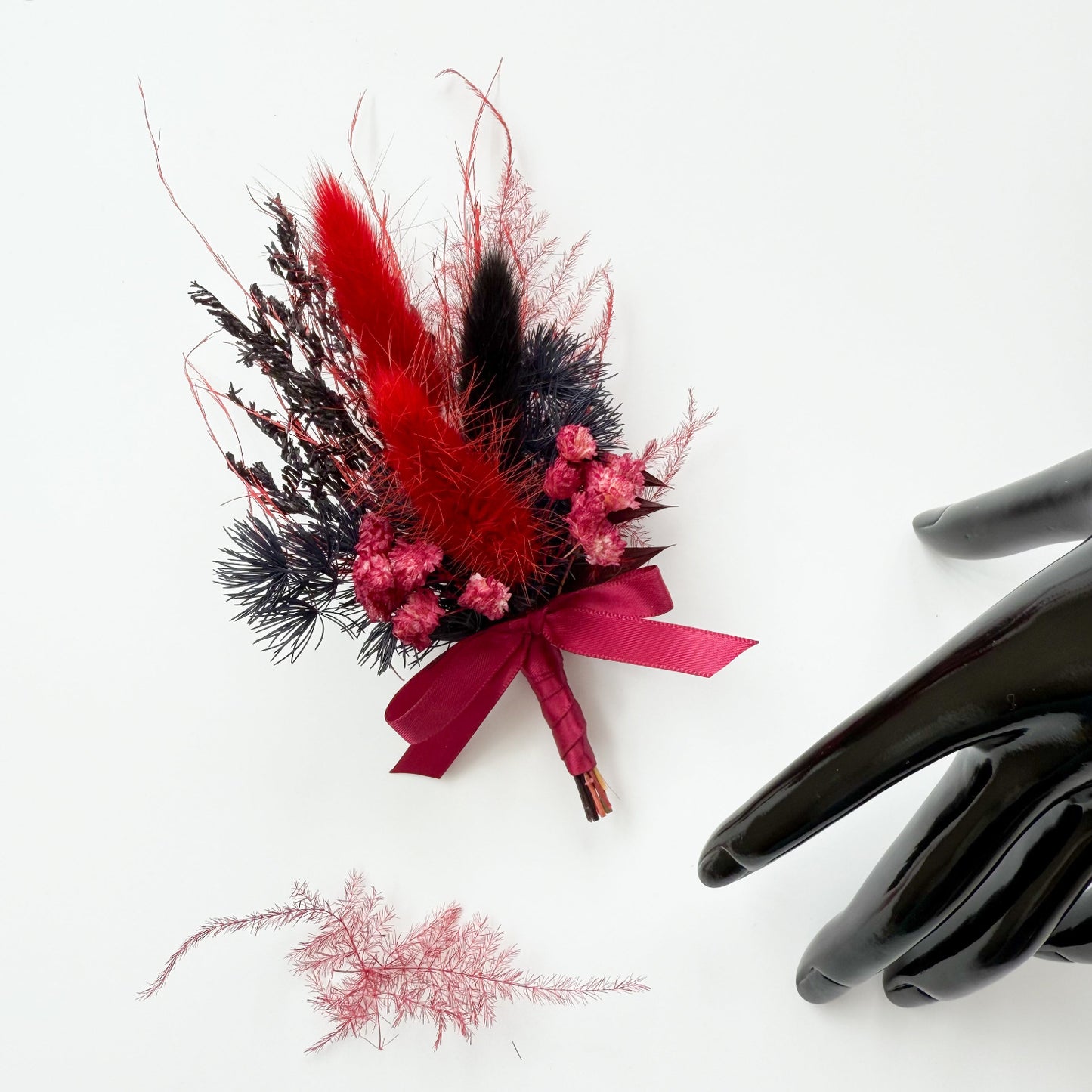 Decorative flower boutonniere with red and black feathers and a pink ribbon, next to a black glove on a white background.