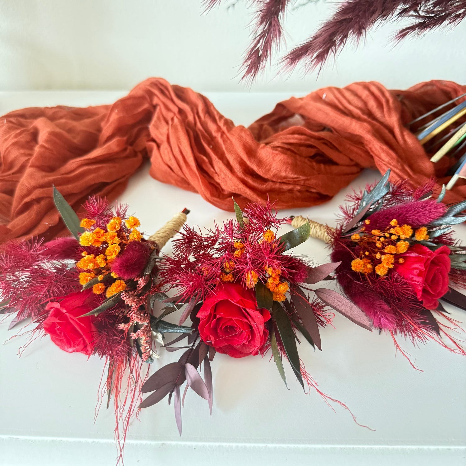 Gothic wedding boutonnieres with burgundy roses, dried flowers, black foliage and burnt orange accents matching a skull wedding bouquet.