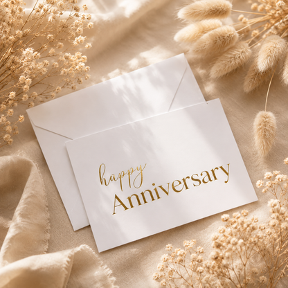 A white anniversary card with elegant gold foil lettering reading 'happy Anniversary' is displayed on a soft beige fabric alongside matching envelopes and dried floral decorations.