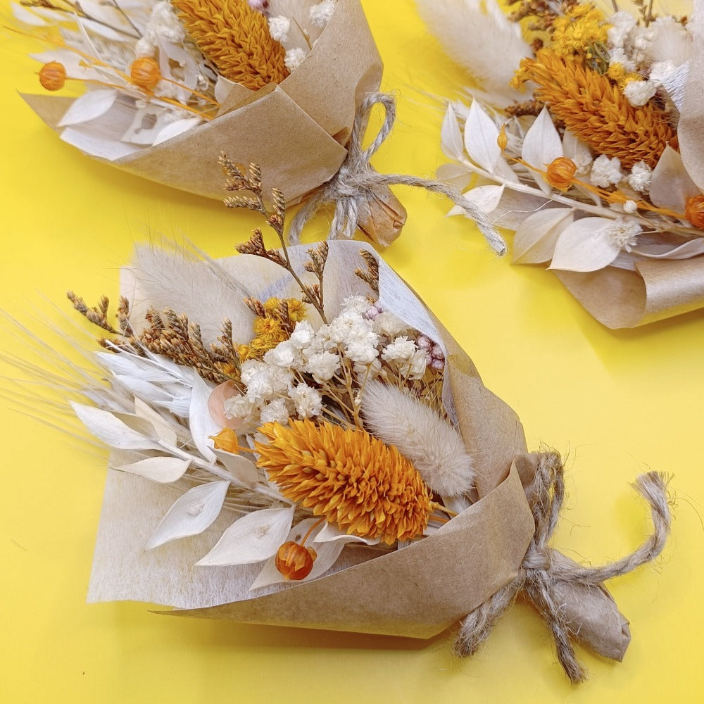 Top-down view of a mini bouquet on a yellow background. Bouquet includes dried yellow protea, billy buttons, white preserved foliage, and neutral dried flowers, tied with rustic twine. Vibrant contrast between the bouquet and the yellow background enhances its cheerful and earthy style. Great for rustic weddings, home decor, or gift sets.