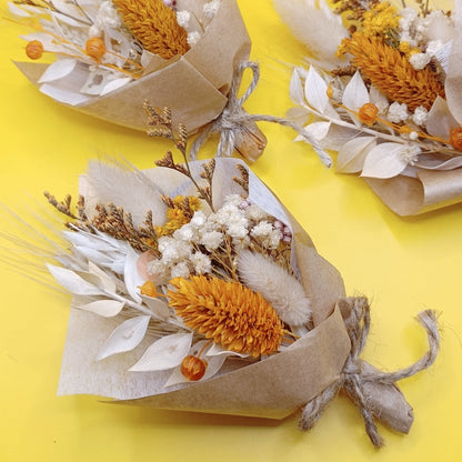Top-down view of a mini bouquet on a yellow background. Bouquet includes dried yellow protea, billy buttons, white preserved foliage, and neutral dried flowers, tied with rustic twine. Vibrant contrast between the bouquet and the yellow background enhances its cheerful and earthy style. Great for rustic weddings, home decor, or gift sets.