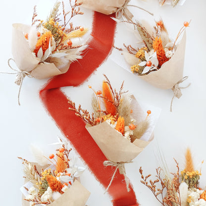 Flat lay of multiple mini dried flower bouquets with warm terracotta, yellow, and cream tones. Arranged on a white background with a bold rust-colored ribbon winding through them. Each bouquet features preserved protea, billy buttons, bunny tails, and kraft paper wrap, perfect for rustic or boho weddings, thank-you gifts, or guest favours. Natural textures and cozy colours create a warm visual appeal.