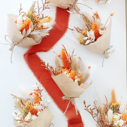 Flat lay of multiple mini dried flower bouquets with warm terracotta, yellow, and cream tones. Arranged on a white background with a bold rust-colored ribbon winding through them. Each bouquet features preserved protea, billy buttons, bunny tails, and kraft paper wrap, perfect for rustic or boho weddings, thank-you gifts, or guest favours. Natural textures and cozy colours create a warm visual appeal.