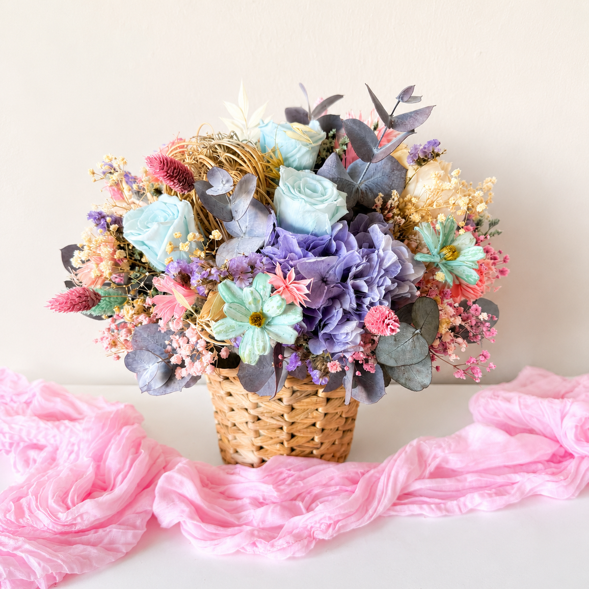 A pastel artificial flower arrangement in a woven basket, featuring light blue roses, lavender hydrangeas, mint daisies, pink filler flowers, and gray-green foliage, set against a plain light beige wall with pink fabric draped in the foreground.
