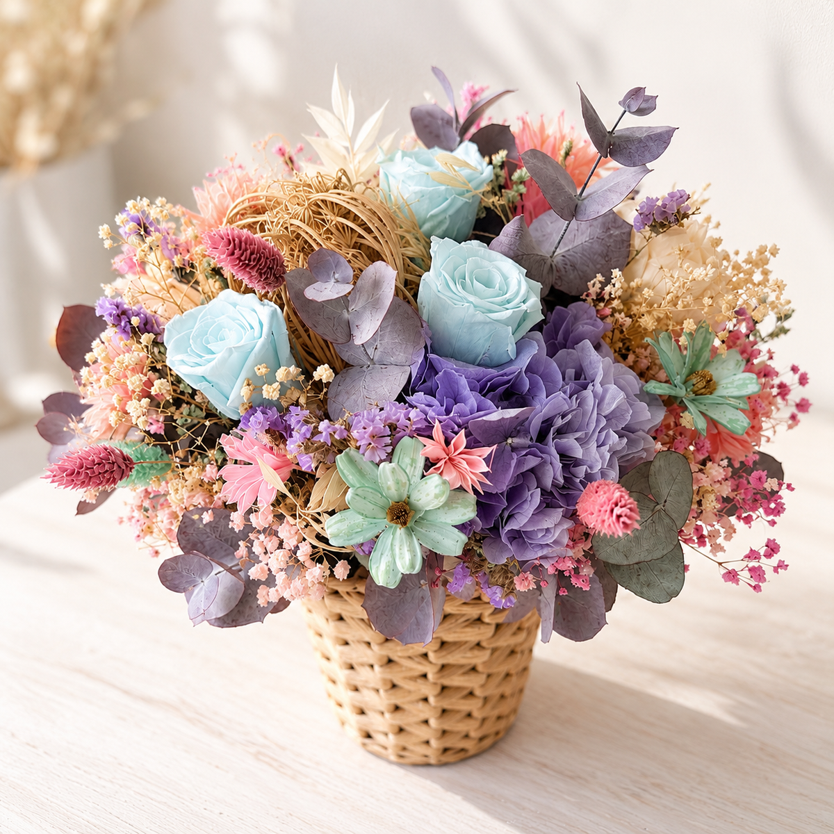 A woven basket filled with a pastel dried flower arrangement, featuring light blue roses, lavender hydrangeas, mint green blossoms, and dusty purple eucalyptus on a light wood surface.