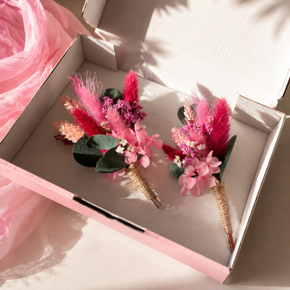Two pink floral boutonniere wristpieces in a light pink gift box, styled with pink tulle fabric on a sunlit beige surface.