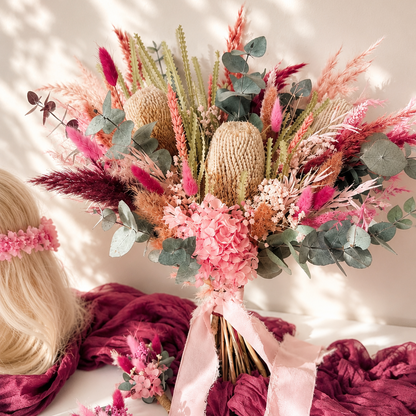 A bohemian-style dried flower bouquet with pink hydrangeas, pink pampas grass, green eucalyptus, and tan kente cloth elements, tied with a light pink ribbon. A blonde model's hair and a matching pink floral crown are visible on the left, with a deep pink textured fabric draped around the base.