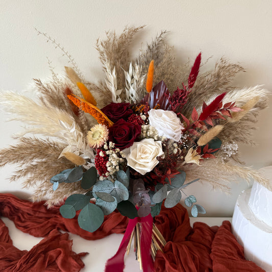 Detailed studio close-up of a luxury dried and preserved floral bridal bouquet, featuring ivory roses, deep crimson amaranthus, feathery pampas grass, and a rich terracotta silk ribbon