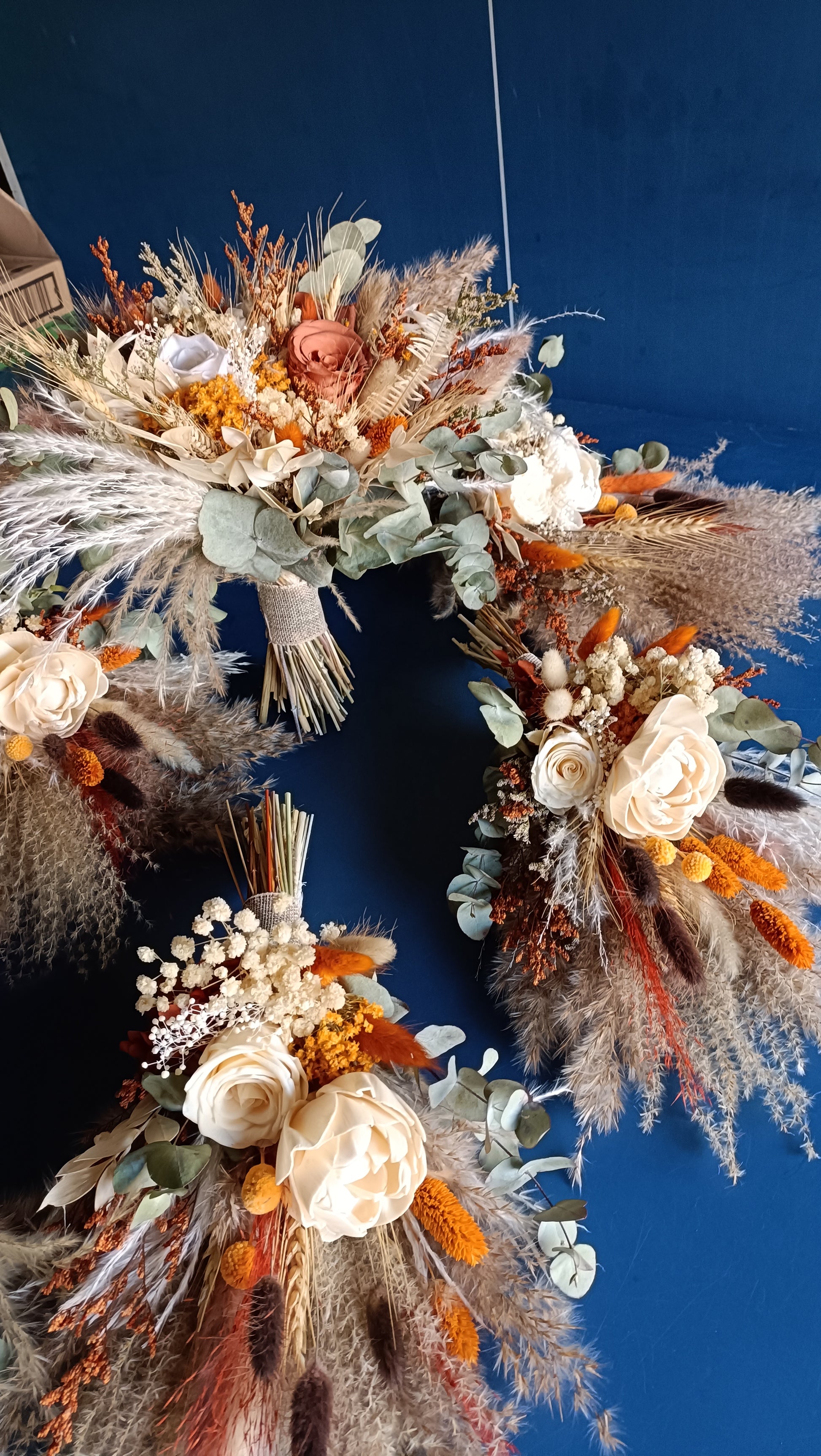 A collection of dried flower wedding bouquets and accessories, showcasing vibrant orange, beige, and green tones. These unique, handcrafted arrangements include preserved roses, eucalyptus, and pampas grass, ideal for boho-chic weddings. Customizable floral designs available.