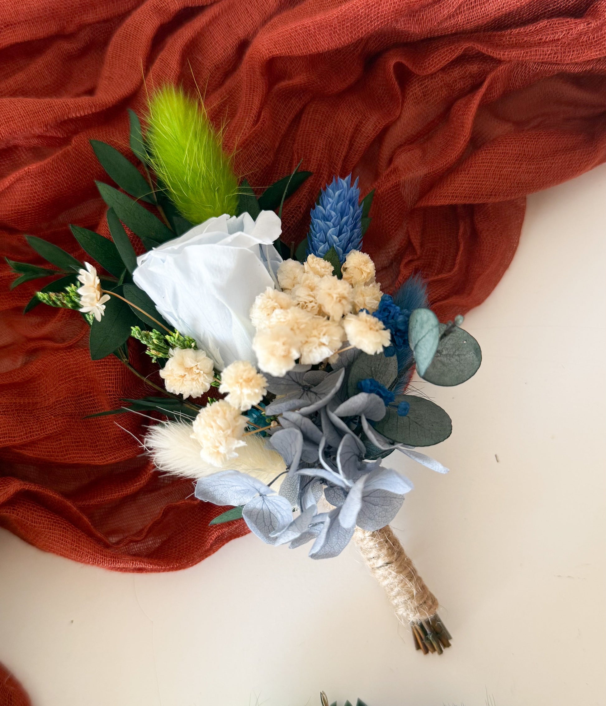 blue boutonniere with dried flowers on a white surface with red fabric in the background