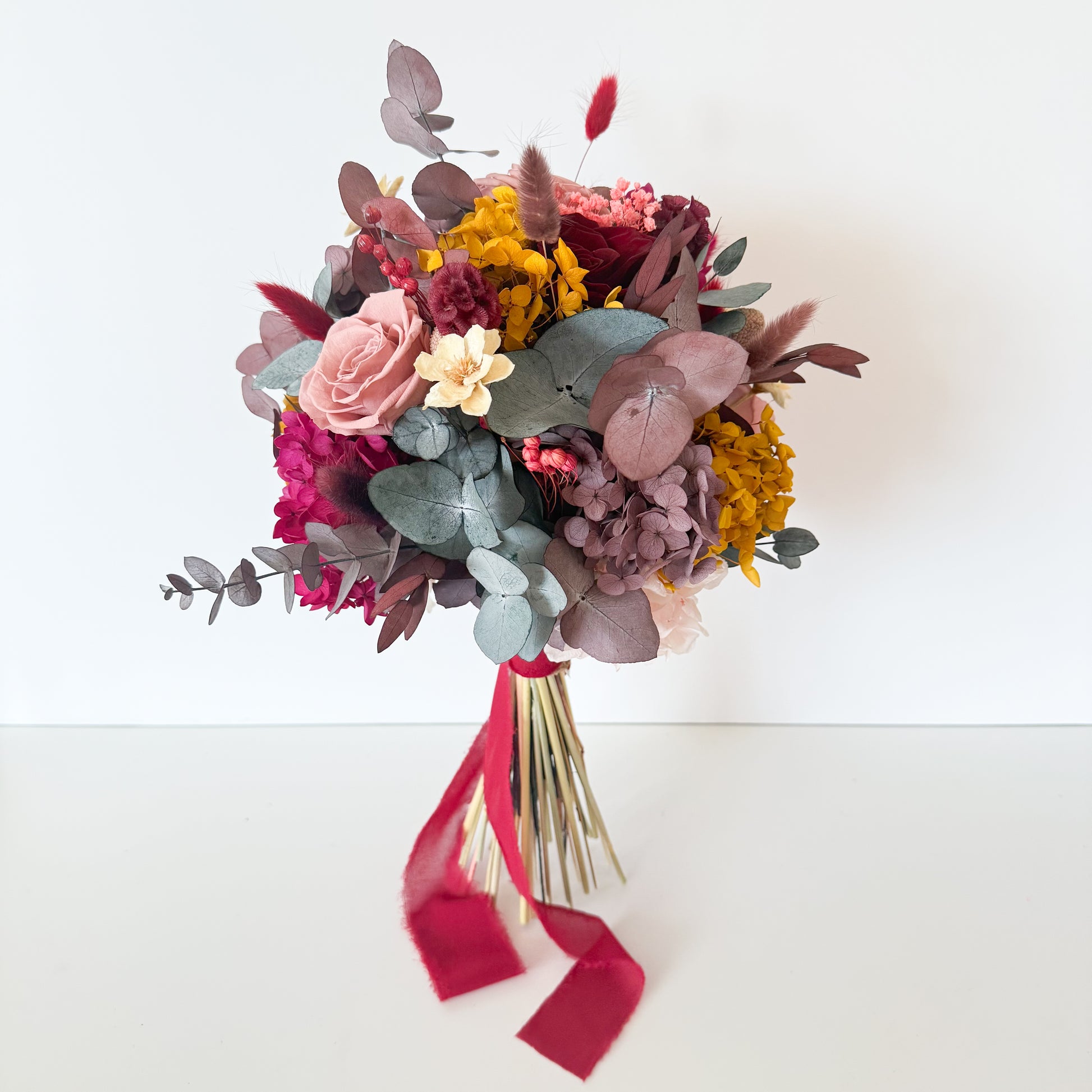 Rustic dried bridal bouquet with dusty pink, cream, and burgundy preserved flowers, tied with a soft red ribbon