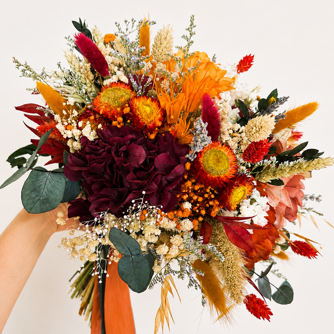 A dried floral bouquet held by a person's hand, featuring orange and red flowers, deep burgundy hydrangea-like blooms, green foliage, and orange ribbon ribbons against a plain off-white background.