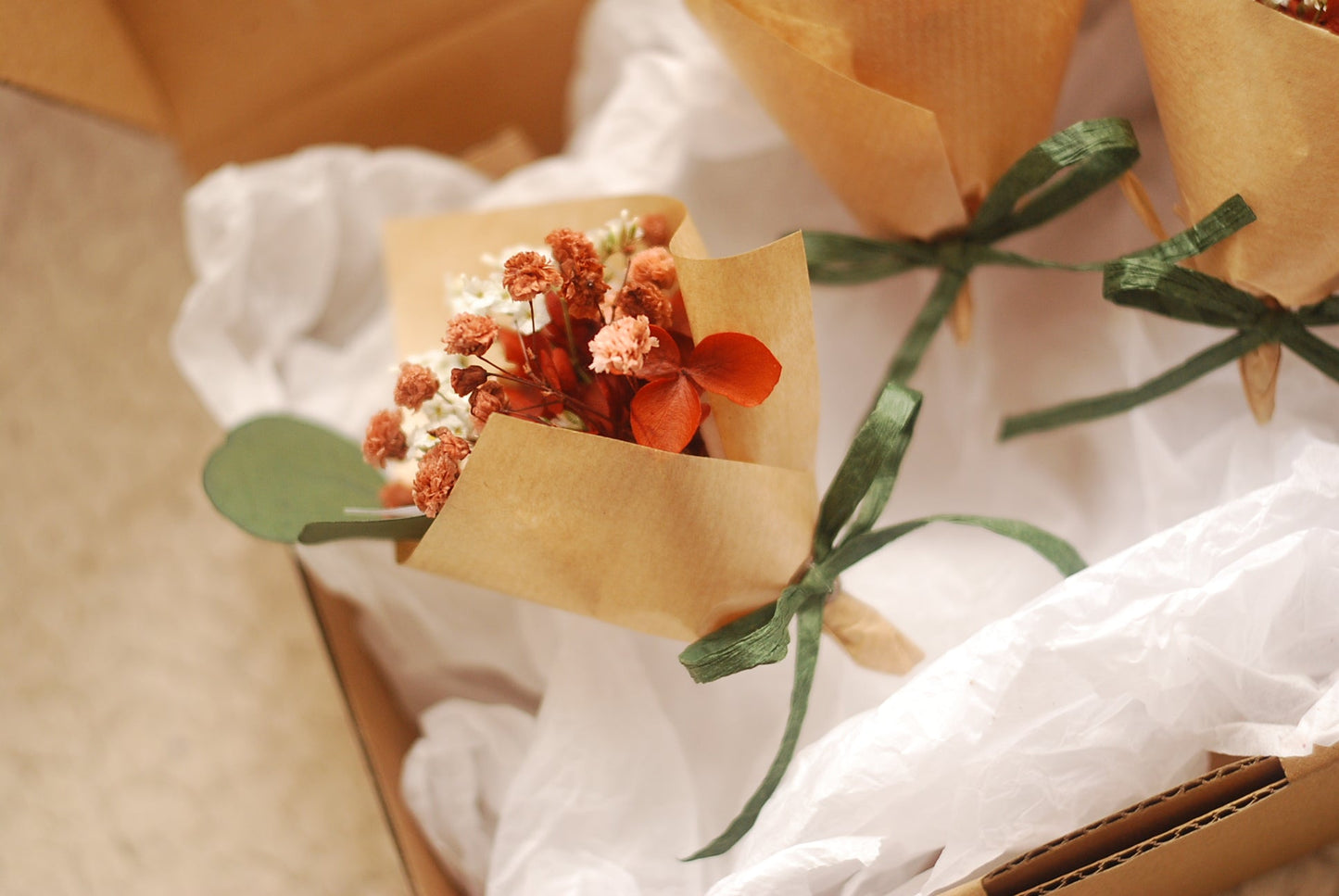 Top view of mini dried bouquet in a box. The bouquet features preserved blooms in warm orange and cream hues, complemented by eucalyptus stems. Packaged with care in kraft paper and tissue wrap, this arrangement suits boho weddings, eco-gifting, and rustic event decor