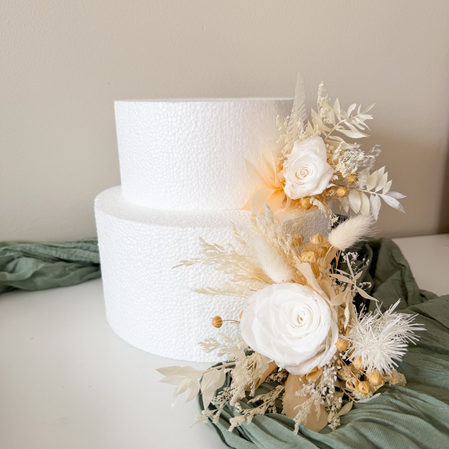 Two-tiered white wedding cake with floral decorations on a light green fabric background.