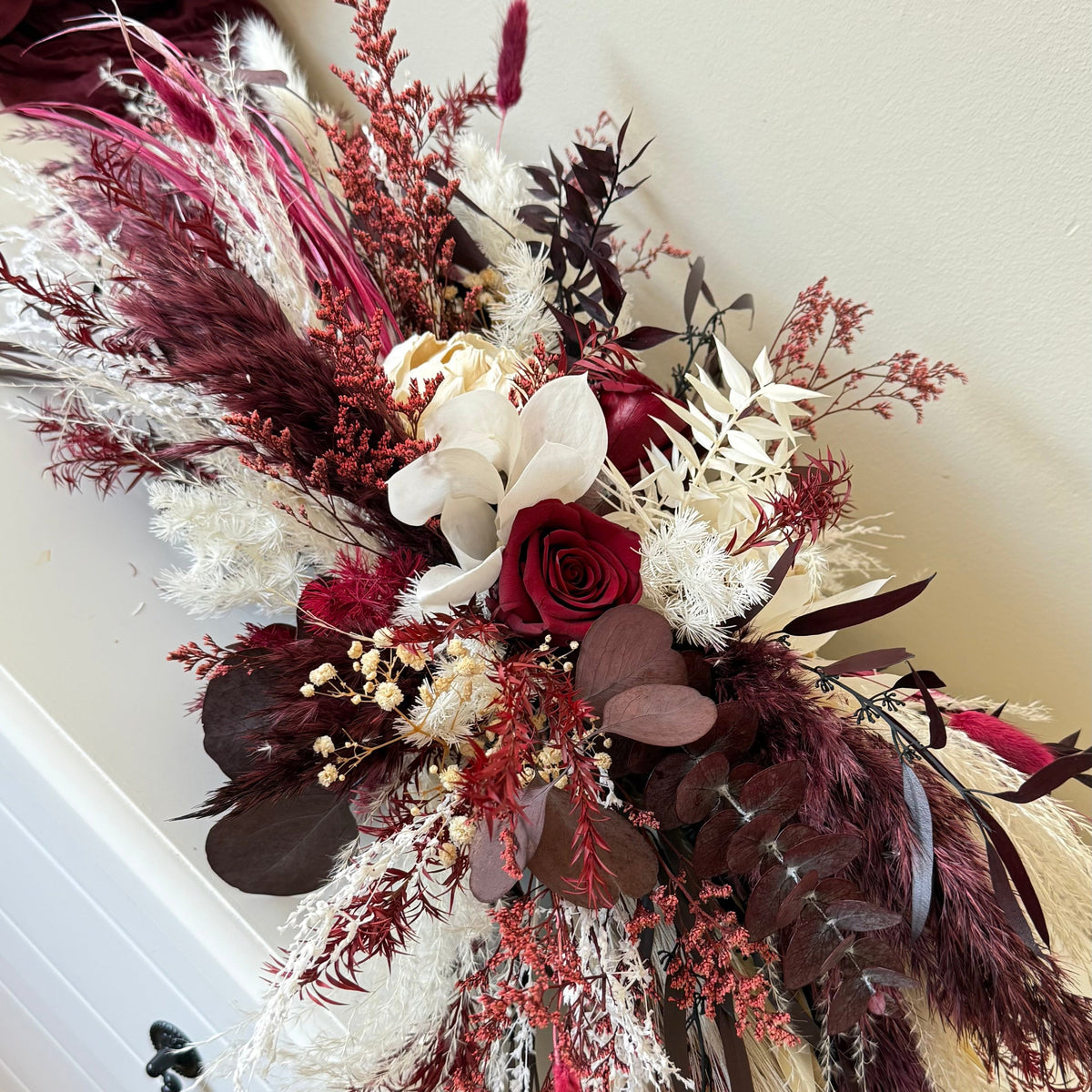 A dried floral arrangement in deep burgundy, cream, and brown tones, styled on a white dresser against a beige wall.