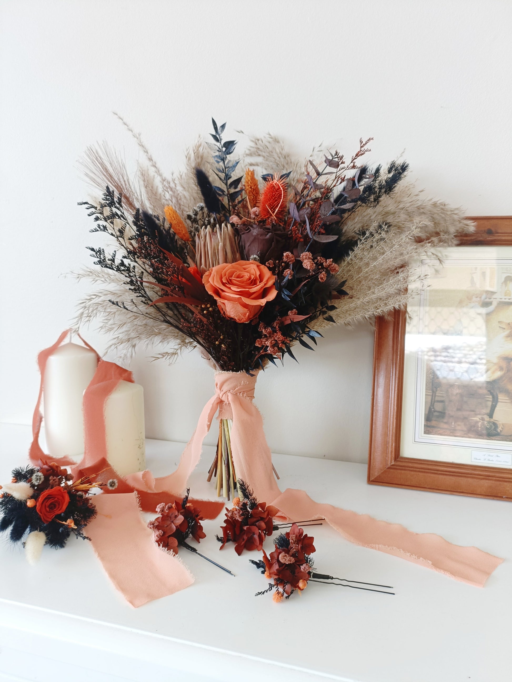 Dried flower bouquet in burnt orange and navy tones with rose centerpiece, paired with coordinating boutonniere and hair accessories on white background.