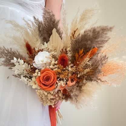 Bouquet of dried flowers and orange roses held by a person wearing a white dress.