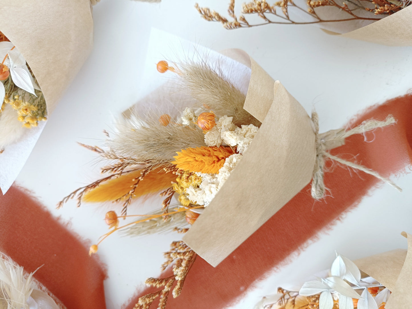 Close-up of a single mini bouquet held in hand, wrapped in kraft paper and twine. The bouquet includes yellow billy buttons, orange bunny tails, preserved protea, and ivory baby's breath. The warm tones and dried textures make it ideal for boho-themed weddings, autumn gifts, or eco-friendly celebrations. The soft light highlights the bouquet's handmade rustic charm.