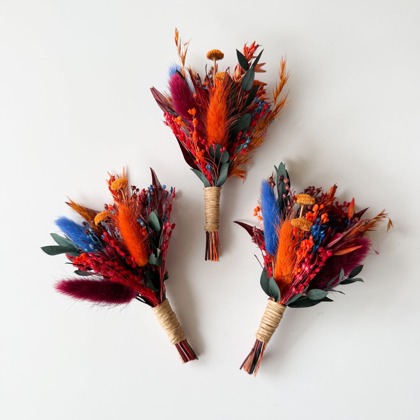 Three vibrant floral boutonnieres with orange, red, and blue flowers on a white background