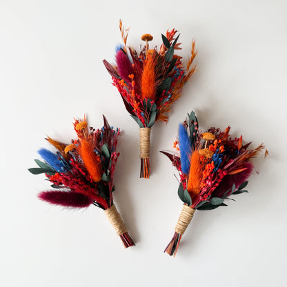 Three vibrant floral boutonnieres with orange, red, and blue flowers on a white background