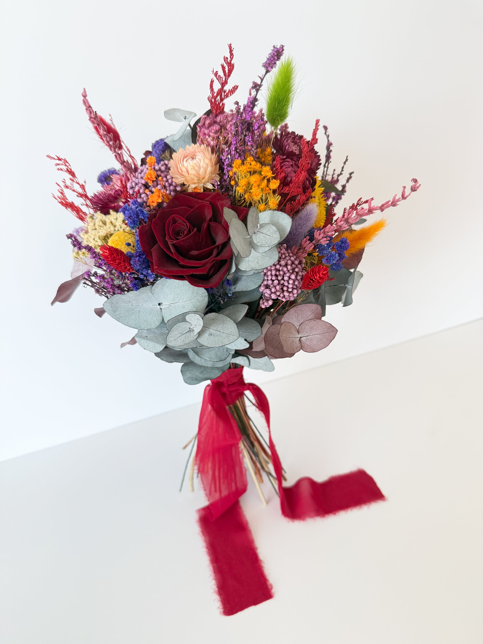 Vibrant dried floral bouquet with yellow billy buttons, burgundy roses, and eucalyptus leaves, finished with red silk ribbon