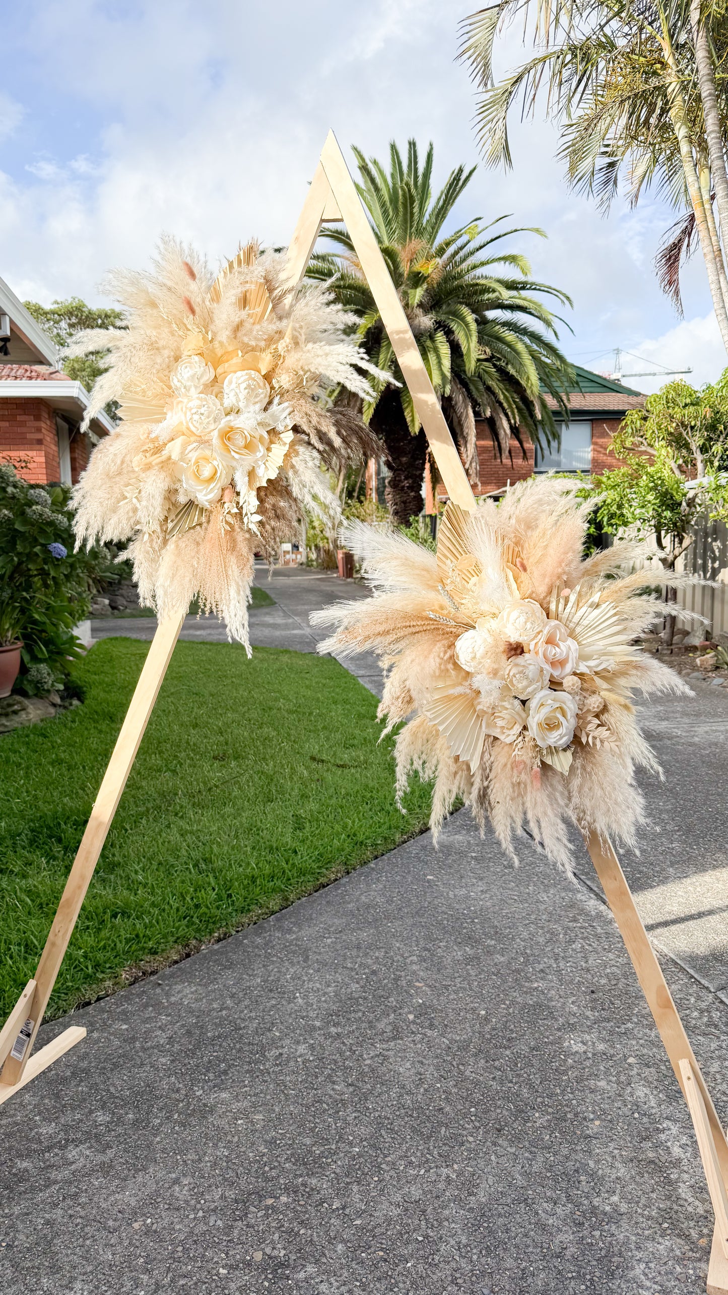 Boho wedding arch flowers with pampas grass, dried palms, and ivory sola peonies and roses arranged on a wooden arch outdoors