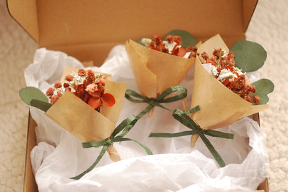 A close-up of a single dried flower bouquet in a box, wrapped in kraft paper with green ribbon and accompanied by eucalyptus leaves. Flowers in shades of terracotta, burnt orange, and white. The soft fabric lining enhances the gift presentation. A thoughtful choice for bridesmaid gifts or sustainable floral favours.

