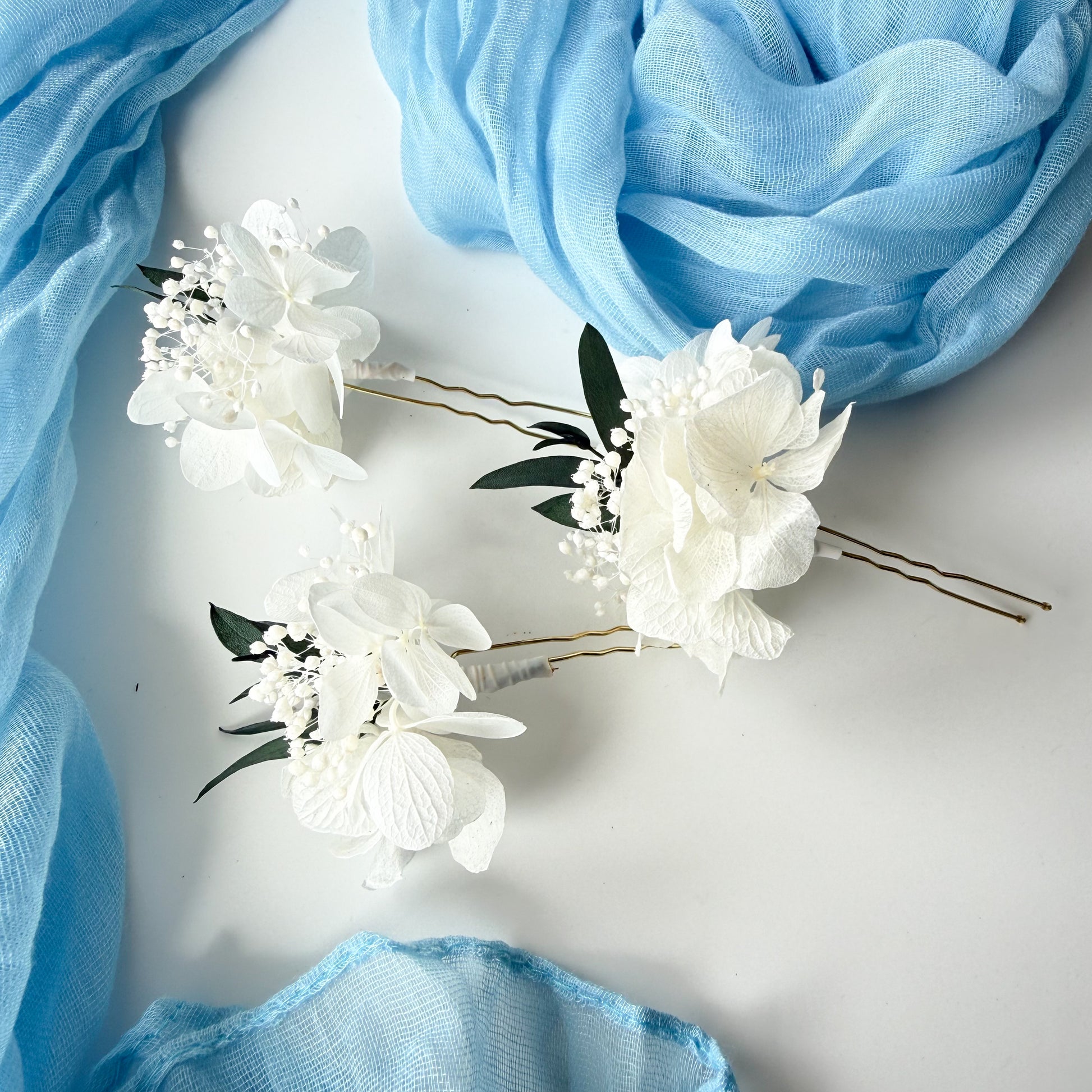 White floral hairpins with green leaves on a light gray background with blue fabric.